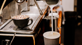 milk being poured into a takeaway coffee cup with cappucino in the background